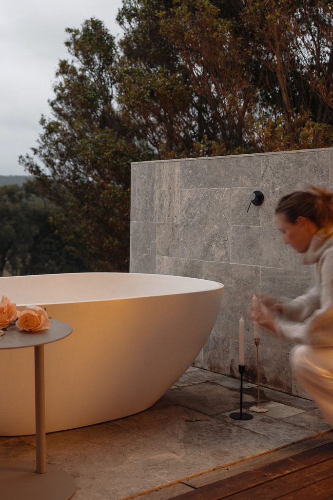 CarrajungEstate_Preview_2026-054 | I do I do Gippsland Outdoor scene with a large white bathtub next to a gray stone wall; a small table with pink roses sits nearby, and a person is blurred at the right edge.