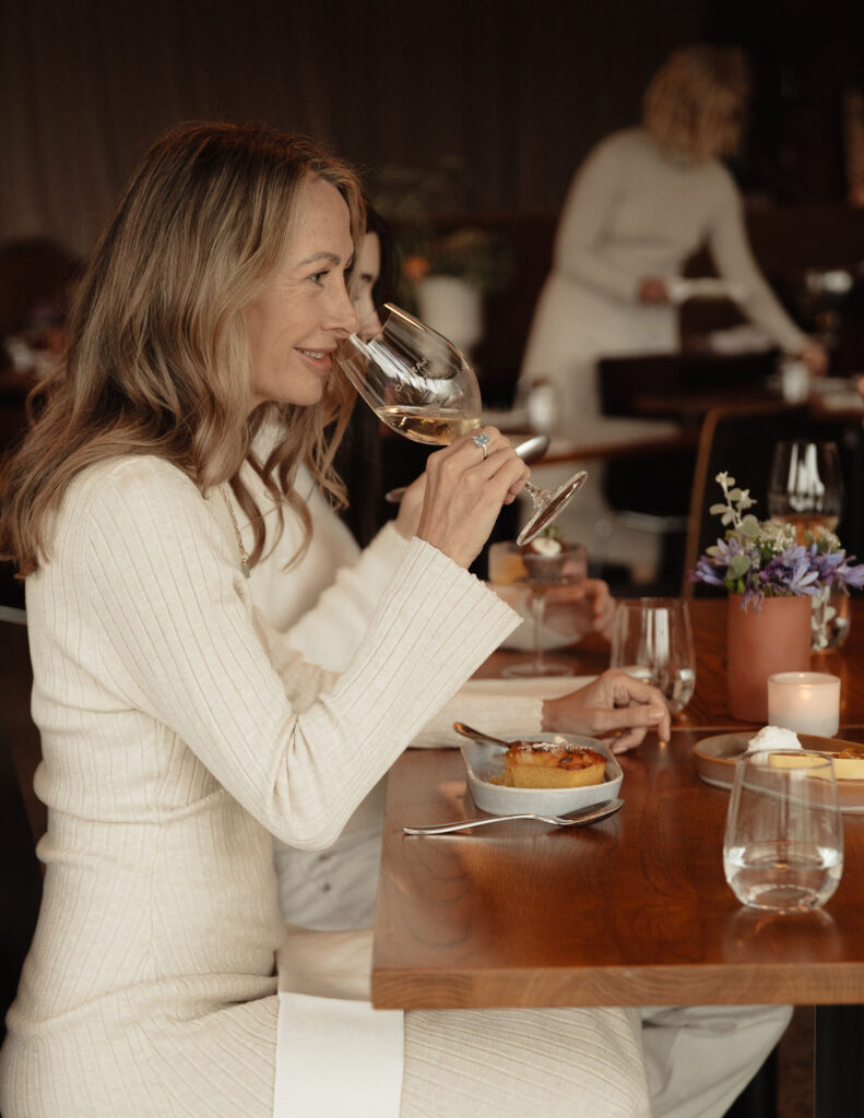 CarrajungEstate_Preview_2026-221 | I do I do Gippsland Woman in a cream sweater sipping white wine at a restaurant table, with desserts and a small candle nearby.