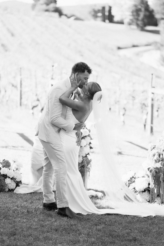 Taylah and Daniel | I do I do Gippsland Bride and groom kiss during an outdoor wedding, embracing in front of floral decorations.