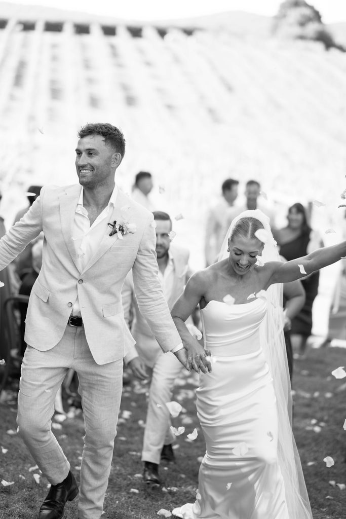 Taylah and Daniel | I do I do Gippsland Bride and groom walking hand in hand through confetti at an outdoor wedding, smiling and joyful as guests celebrate behind them (black and white)