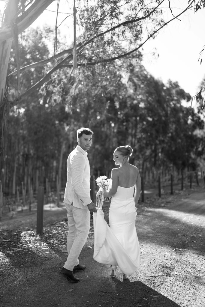Taylah and Daniel | I do I do Gippsland Bride in a white wedding dress and a light-colored suit groom walk hand in hand along a tree-lined path; she holds a bouquet.