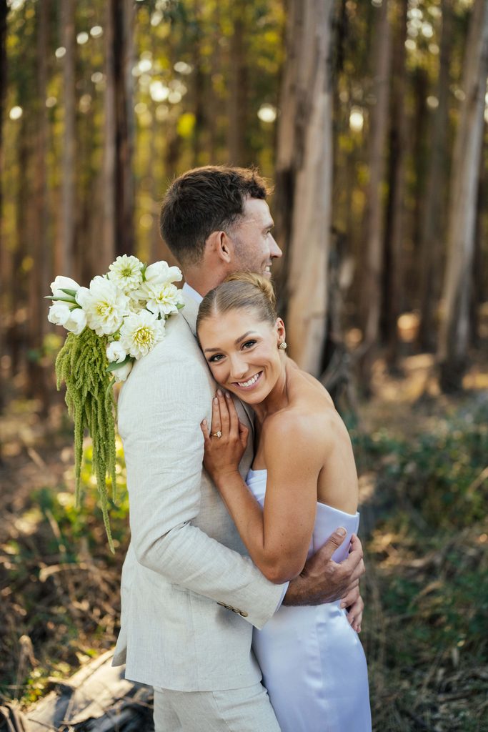 Taylah and Daniel | I do I do Gippsland A couple hugs in a sunny forest; the woman in a white dress rests her head on the man's chest, smiling with a bouquet visible on his back at Carrajung Estate, your Gippsland Wedding Venue.