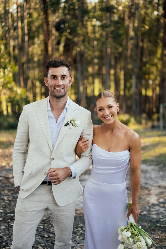 Taylah and Daniel | I do I do Gippsland Smiling bride and groom posing together on a forest path; the groom wears a light tan suit and the bride a strapless white gown, holding a white bouquet.